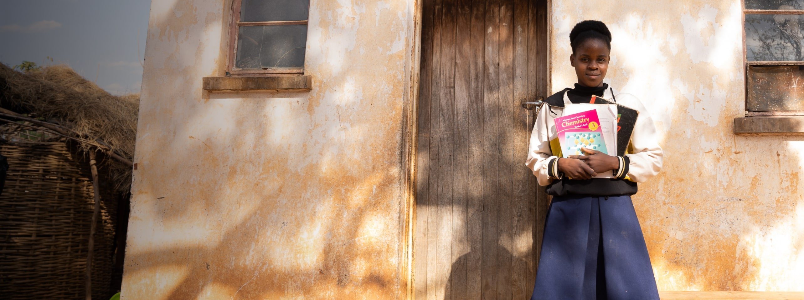 A student holding chemistry text books, standing in front of a rustic home
