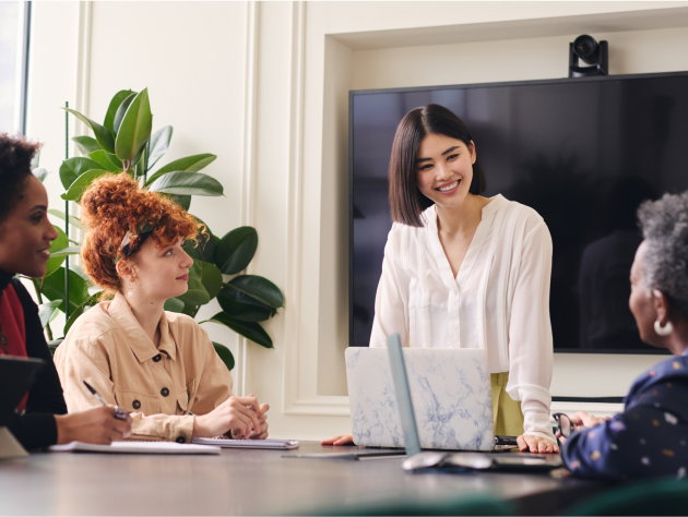 Three women of different ages, one Asian woman with black hair and a white shirt smiling at each other in front of their laptops sitting around a conference table in an office room. A big screen TV is mounted on the wall in a room with a modern design, simple color scheme, and natural light. The photography style is professional.